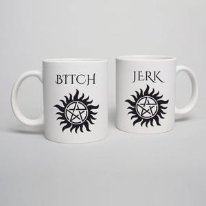 May include: Two white ceramic mugs. One mug has the word "BITCH" above a black sunburst design with a pentagram. The other mug has the word "JERK" above a similar black sunburst design.