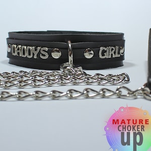 May include: Black leather choker with silver studs and the words "Daddy's Girl" on it. A silver chain is attached to the choker. The choker is on a white background.