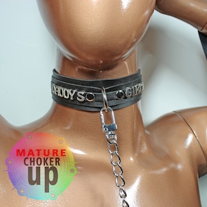May include: Black leather choker with silver metal accents and the words "Bad Boys" and "Girl" on it. The choker has a silver metal chain attached to it.