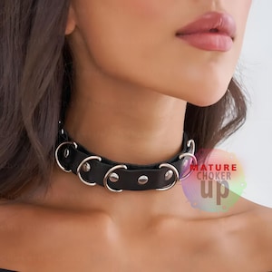 Op de afbeelding: Zwarte leren choker ketting met zilverkleurige metalen ringen en studs. De choker wordt om de nek gedragen. De tekst "MATURE CHOKER up" is zichtbaar in de afbeelding.