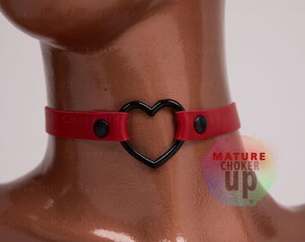 May include: Red leather choker with a black heart-shaped metal ring. The choker has two black snap closures. The text "MATURE CHOKER up" is visible in the background.