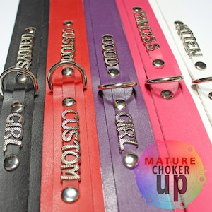 May include: Five leather chokers with metal rings and studs. The chokers are black, red, purple, pink, and white. Each choker has a different word embossed on it: "Bad Girl", "Custom", "Good Girl", "Princess", and "Milen".