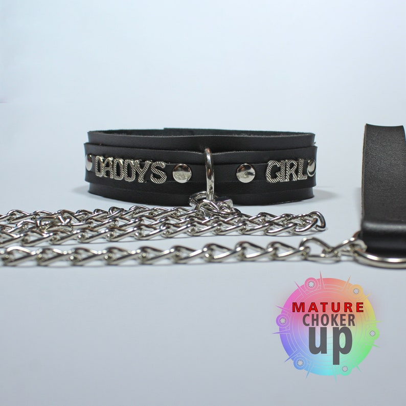 May include: Black leather choker with silver studs and the words "Daddy's Girl" on it. A silver chain is attached to the choker. The choker is on a white background.