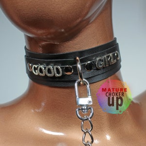 May include: Black leather choker with silver metal accents and the words "GOOD GIRL" in raised letters. The choker has a silver metal clasp and a chain attached.