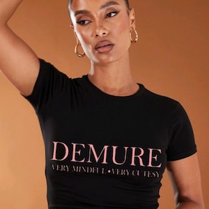 May include: Black crew neck t-shirt with the word "DEMURE" in pink script across the chest. Below the word "DEMURE" are the words "VERY MINDFUL • VERY CUTESTY" in a smaller pink font.