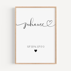 May include: A framed print featuring the word "zuhause" in black script, with a heart symbol. Beneath, coordinates and a black heart are shown. The frame is a light wood colour.