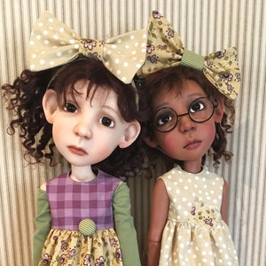 May include: Two handmade dolls stand side-by-side, each with a unique style. One doll wears a purple plaid dress with a floral skirt, green leggings, and cream-colored shoes. The other doll has a polka-dot dress, brown leggings, and tan boots.