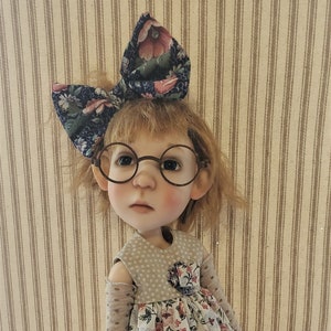 May include: A doll with brown hair wearing a floral dress, a blue and white bow, and round glasses. The doll is wearing pink shoes and has a light brown complexion.