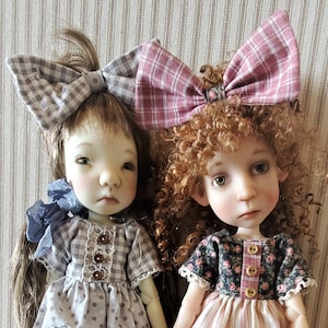 Puede incluir: Dos muñecas de porcelana que llevan vestidos y zapatos. La muñeca de la izquierda tiene el pelo castaño con un lazo a cuadros gris y blanco. La muñeca de la derecha tiene el pelo rojo rizado con un lazo a cuadros rosa y blanco.