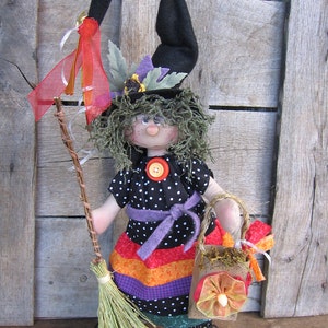 Puede incluir: Una muñeca de bruja hecha a mano con un sombrero negro, un vestido a rayas morado y naranja y una escoba. La muñeca lleva una pequeña bolsa marrón con un diseño floral.