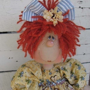 Könnte beinhalten: Eine handgemachte Stoffpuppe mit roten Wollhaaren, einer blau-weiß gestreiften Schleife und einer gelben Blumenverzierung. Die Puppe trägt ein gelbes Kleid mit Blumenmuster und einen herzförmigen Knopf.