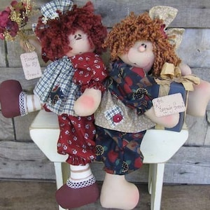 May include: Two handmade rag dolls sitting on a white stool. The doll on the left is wearing a plaid dress with red and white polka dot pants and red shoes. The doll on the right is wearing a blue and red dress with a brown and white patchwork bodice and brown shoes. The doll on the right is holding a blue gift box with a gold ribbon and a tag that says "My Special Friend".