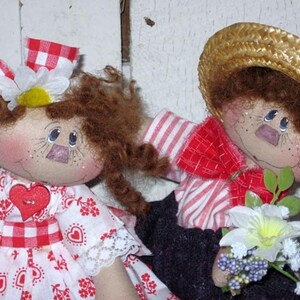 May include: Two handmade fabric dolls. One doll wears a red and white checkered dress with a heart-shaped button and a matching bow. The other doll wears a straw hat, red and white striped shirt, and holds a bouquet of flowers. Both dolls have freckles and painted features.