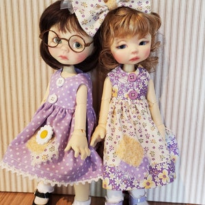 Puede incluir: Dos muñecas con vestidos morados con estampados florales y lunares blancos. Las muñecas tienen el pelo rubio y castaño y llevan zapatos negros y morados.