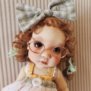 May include: A doll with curly red hair wearing a green and white plaid bow, round glasses, and a yellow and white floral dress with a white lace trim.