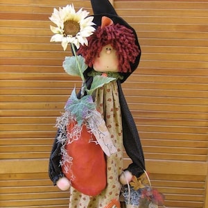 May include: A handmade rag doll with red yarn hair wearing a black witch hat and a brown and green dress. The doll is holding a sunflower and a brown paper bag with fall leaves.