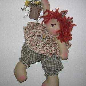 Puede incluir: Una muñeca de trapo hecha a mano con cabello de hilo rojo, un vestido con estampado floral y pantalones a cuadros. La muñeca está colgada en una pared, con una pequeña maceta de flores encima.