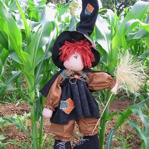 May include: A handmade witch doll with red hair, wearing a black dress, a black witch hat, and black boots. The doll is holding a broom and is standing in a field of corn.