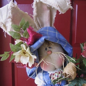 May include: A handmade bunny doll wearing a blue plaid dress and a white hat with frayed edges. The doll is holding a bouquet of flowers and has a sweet, whimsical expression.