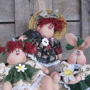 Op de afbeelding: Drie handgemaakte stoffen poppen, twee met rood haar en een met konijnenoren. De poppen dragen bloemenjurken met witte kant en zitten op een houten oppervlak.