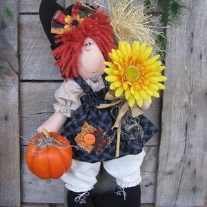 May include: A handmade rag doll dressed in a black and blue patchwork dress with a white apron and black shoes. The doll is holding a small orange pumpkin and a yellow sunflower. The doll is standing in front of a wooden background.
