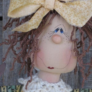 以下が含まれることがあります： 茶色の毛糸の髪、黄色の花柄の布のリボン、白い編み物の襟が付いた手作りのかやの人形。人形は青い目と赤い口が描かれた顔をしています。