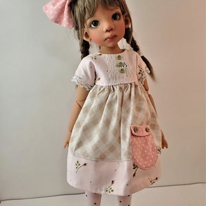 May include: A doll wearing a pink and brown dress with a white lace trim, a pink polka dot bow in her hair, and brown shoes. The doll has brown hair and brown eyes.