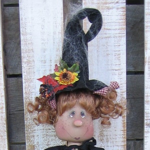 May include: A handmade Halloween witch doll with a black hat, adorned with a sunflower, red leaves, and a spider. The doll has red curly hair, rosy cheeks, and a black outfit. The background is a rustic white wooden surface.