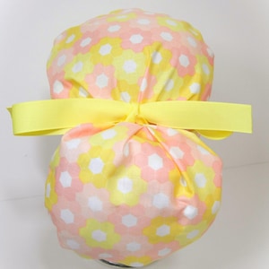 May include: A floral patterned scrub cap in yellow, pink, and white, tied with a bright yellow ribbon. The cap features a repeating hexagonal flower design. The fabric is soft and the bow is centered.