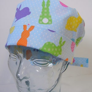 May include: A light blue scrub hat with a white polka dot pattern and colorful bunny rabbit illustrations. The hat has a tie on the side.