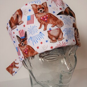 Puede incluir: Un gorro de quirófano blanco con un patrón repetido de perros, fuegos artificiales y estrellas. Los perros llevan bandanas rojas, blancas y azules. El gorro tiene una corbata en el lateral.