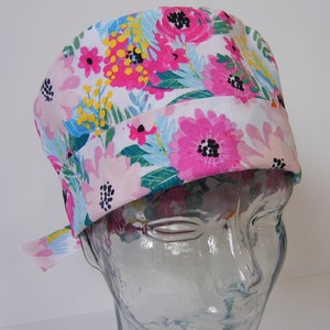 Pode incluir: Um gorro de cirurgia branco com estampa floral rosa, azul e amarela. O gorro tem um laço lateral.