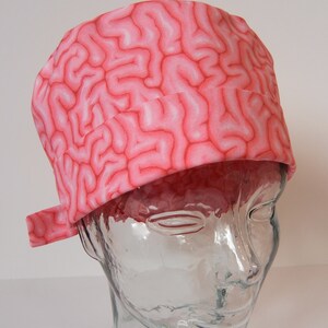 May include: A pink and white patterned surgical scrub hat with a brain design. The hat is on a clear glass head form.