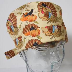 Pode incluir: Um gorro de cirurgia bege com estampa de peru de Ação de Graças. O gorro apresenta vários perus com penas vermelhas, azuis e marrons, junto com abóboras e um pequeno pássaro preto.