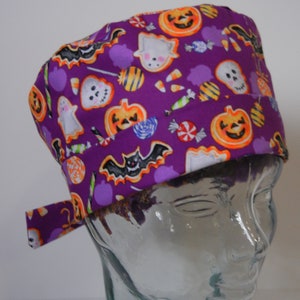 Può includere: Un cappello chirurgico viola di Halloween con un motivo ripetuto di zucche, fantasmi, pipistrelli e caramelle. Il cappello è in tessuto e ha un laccio sul retro. Il design include elementi arancioni, bianchi e neri.