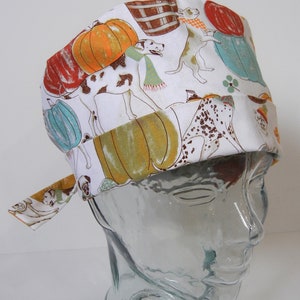 May include: White scrub hat with a colorful print of pumpkins, dogs, and barrels. The print is in shades of orange, brown, white, and blue.