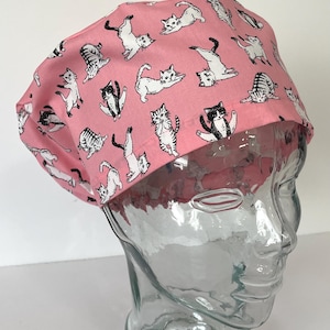 May include: A pink scrub cap featuring a repeating pattern of various black, white, and gray cats in different playful poses. The fabric head covering has a rounded top and a gathered back section, designed to fit securely around the head.