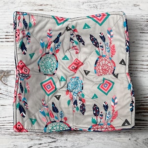 Könnte beinhalten: Quadratische Schüssel-Hülle aus grauem Stoff mit einem Muster aus Traumfängern und Federn in Türkis, Rosa und Marineblau. Das Design umfasst geometrische Formen. Die Hülle hat einen dunkelblauen Rand und ist gesteppt.