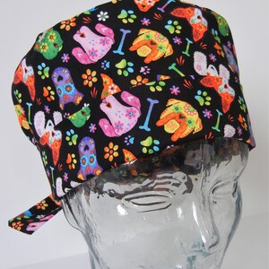 Op de afbeelding: Zwarte scrub-muts met een kleurrijke print van suiker schedel honden, voetafdrukken en bloemen.