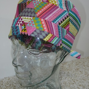 Può includere: Un copricapo chirurgico colorato con un motivo geometrico in tonalità rosa, blu, verde e marrone. Il copricapo è in tessuto e ha un laccio sul retro.