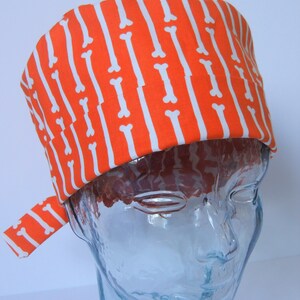 Può includere: Cappello chirurgico arancione e bianco con motivo a osso. Il cappello è su una testa di manichino in vetro trasparente.