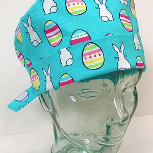 May include: A turquoise blue scrub hat with a repeating pattern of white bunnies and colorful Easter eggs.