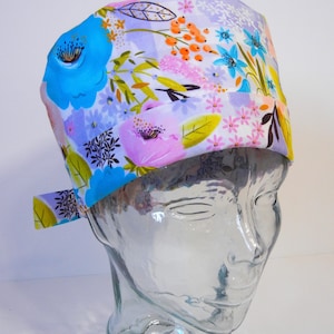 May include: A floral patterned scrub cap featuring a vibrant mix of blue, purple, pink, and orange flowers on a white background. The cap is displayed on a clear glass mannequin head, showcasing its design and fit. The cap has a tie in the back.