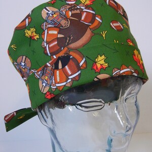 Pode incluir: Um gorro de cirurgia verde com um padrão de perus jogando futebol e folhas de outono. O gorro tem um laço na parte de trás.