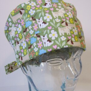 Puede incluir: Un gorro de quirófano verde con un patrón de perros, gatos, mariposas y flores. El gorro tiene un lazo en el lateral.