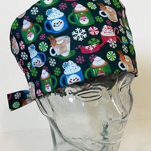 Może przedstawiać: Zielony czepek chirurgiczny z świątecznym wzorem przedstawiającym kubki z gorącą czekoladą, laski cukrowe i płatki śniegu.