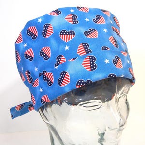 Könnte beinhalten: Eine blaue OP-Haube mit einem Muster aus rot-weiß-blauen Herzen und Sternen. Die Herzen sind mit Streifen und Sternen gestaltet, die an die amerikanische Flagge erinnern. Die Haube hat hinten einen Bindeband zur Anpassung.