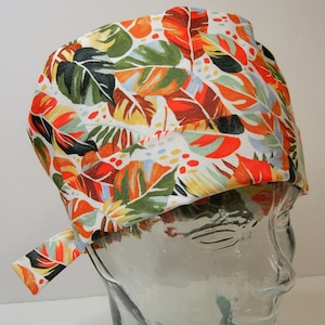 Pode incluir: Um gorro de cirurgia branco com uma estampa tropical de folhas coloridas em tons de laranja, verde e marrom. O gorro tem um laço na parte de trás.