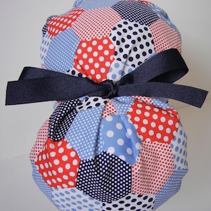 May include: A blue, red, and white fabric pouch with a navy blue ribbon tied around it. The pouch is made of patchwork fabric with polka dots and geometric shapes.