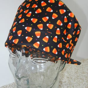 Può includere: Un cappello in tessuto nero con un motivo di mais dolce arancione e bianco e piccoli punti viola e verdi. Il cappello è su una testa di manichino di vetro.